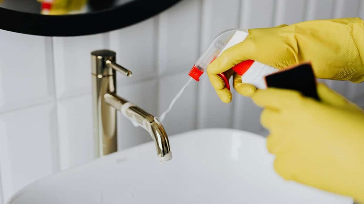 From above anonymous housekeeper in yellow rubber gloves spraying cleaner to chrome water faucet in bathroom