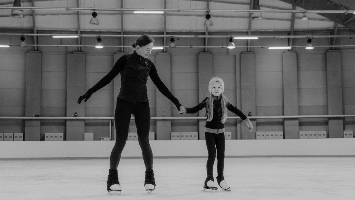 A mother teaching her daughter figure skating in an indoor ice arena.