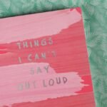 Pink notebook with the text 'Things I Can't Say Out Loud' on green leafy surface.