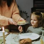 A mother serves dinner to her young daughter at a warmly lit family table, creating a cozy atmosphere.