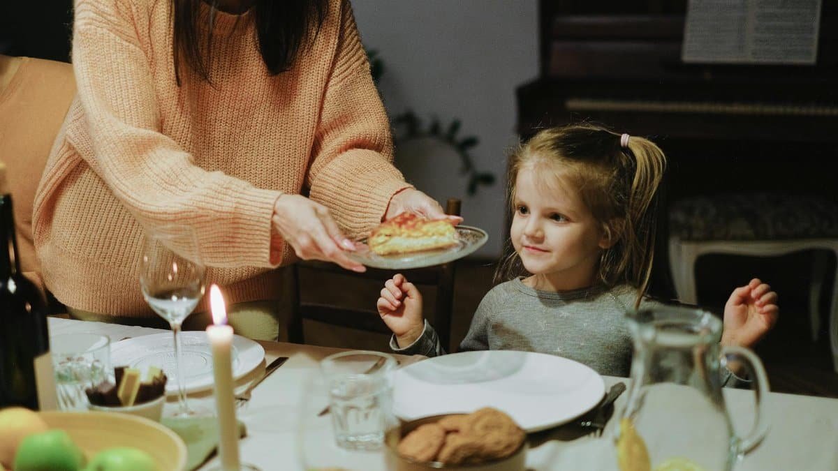 A mother serves dinner to her young daughter at a warmly lit family table, creating a cozy atmosphere.