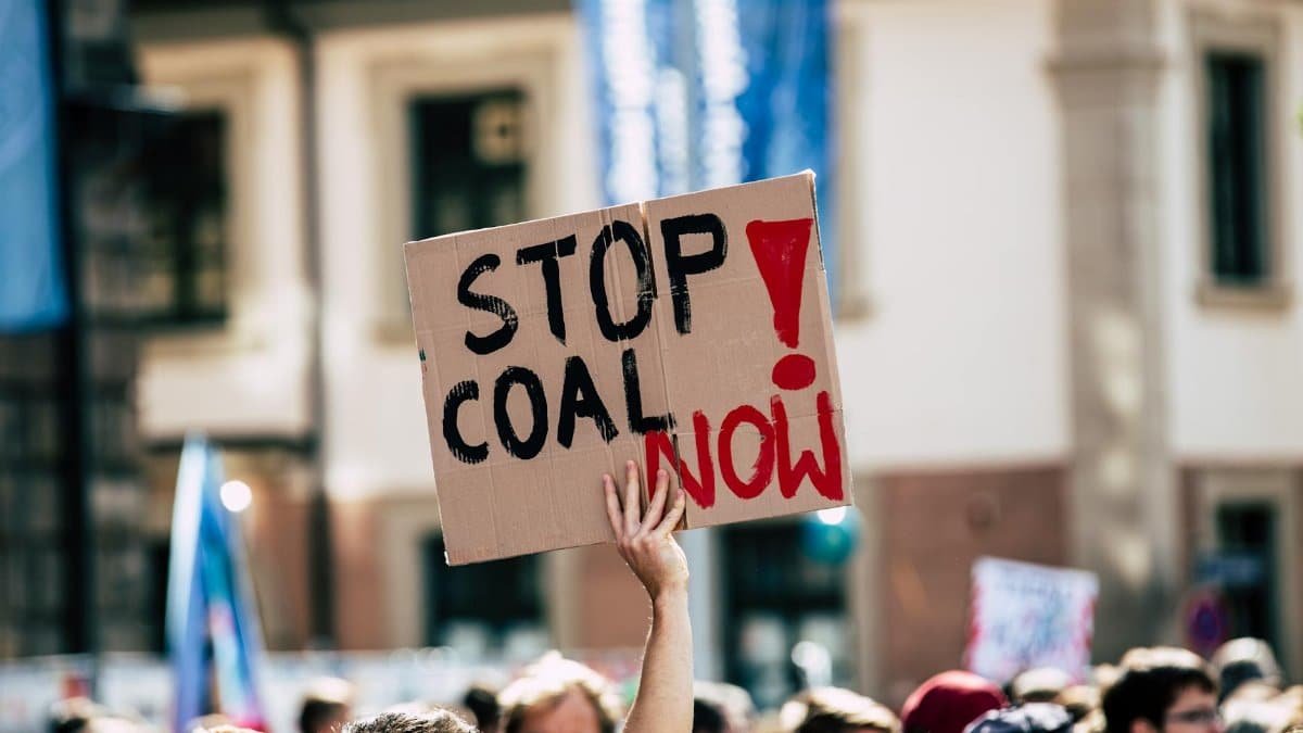 A crowd gathers for an outdoor protest with a sign reading 'Stop Coal Now'.