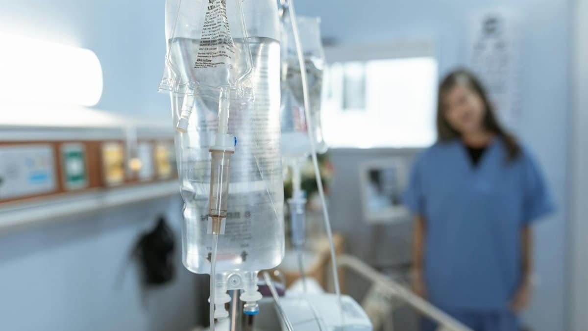 Close-up of an IV drip in a hospital with a blurred nurse in the background.