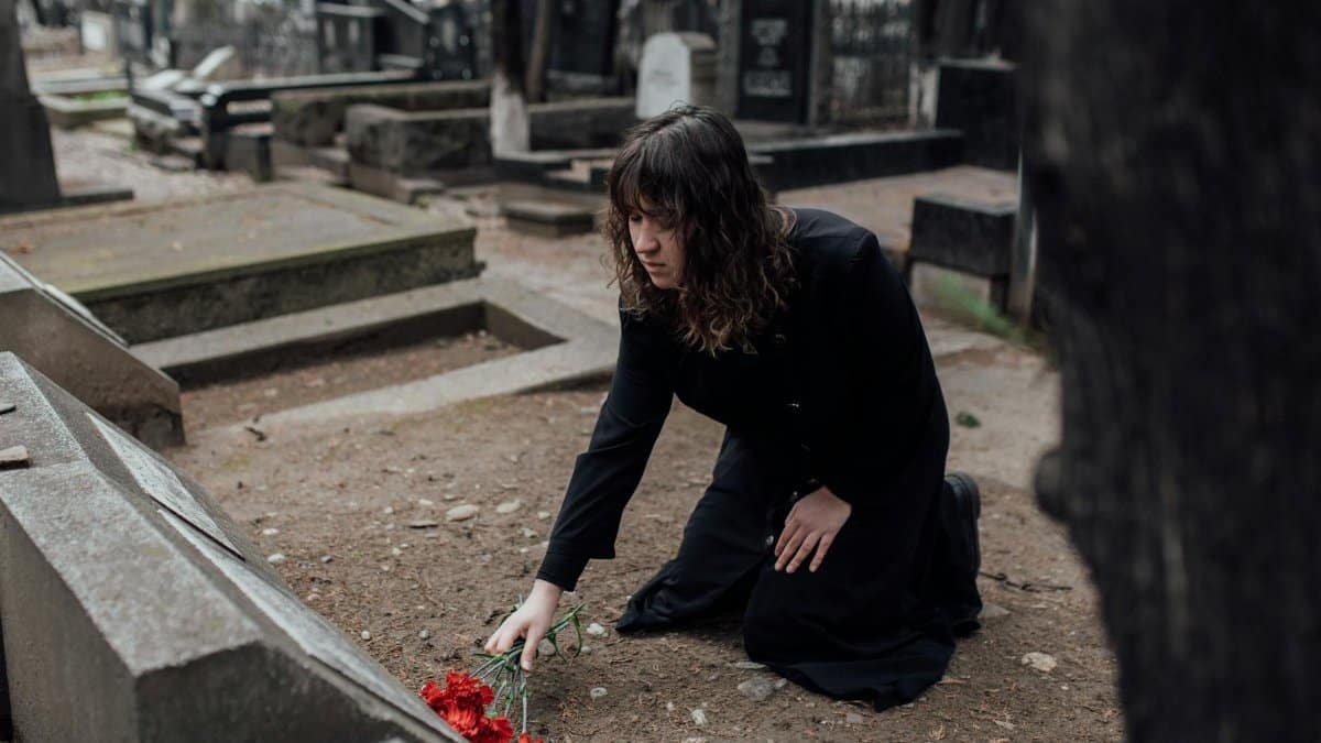 A woman in black kneels in a cemetery placing flowers on a grave.