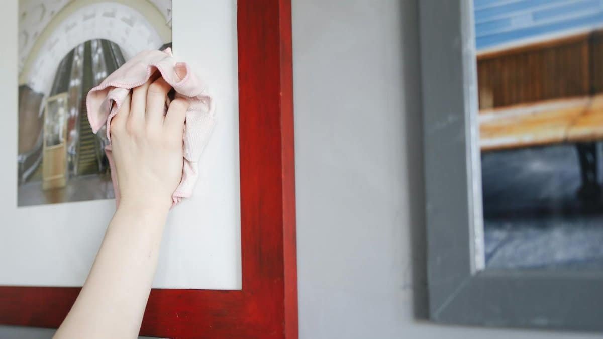 A hand using a cloth to clean a picture frame indoors. Detailed shot of housekeeping.