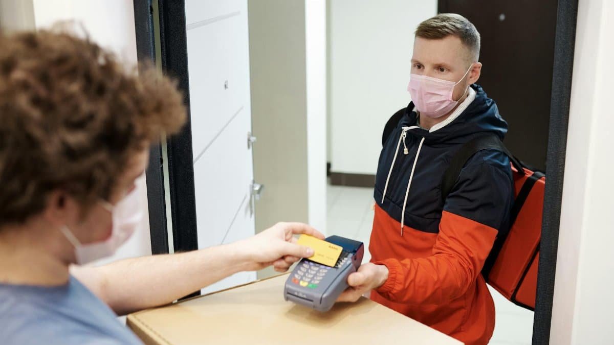 Masked deliveryman receiving payment for pizza delivery, highlighting safety measures.