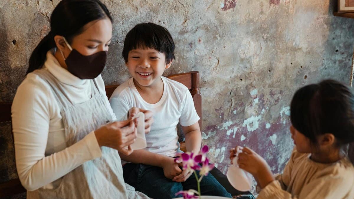Optimistic Asian mother in face mask putting respirators on smiling son and daughter while spending time together in rural house against shabby wall