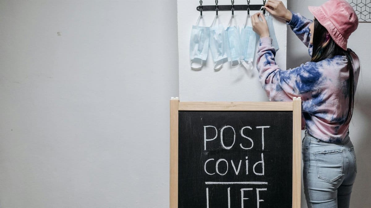 A person hangs masks near a chalkboard with 'Post Covid Life' written, symbolizing the transition to normalcy.