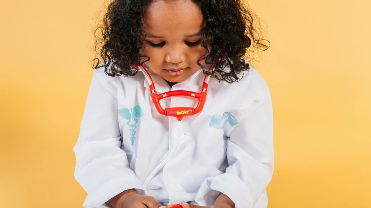 Tranquil little African American girl with curly hair in medical costume and stethoscope looking down against yellow background in studio