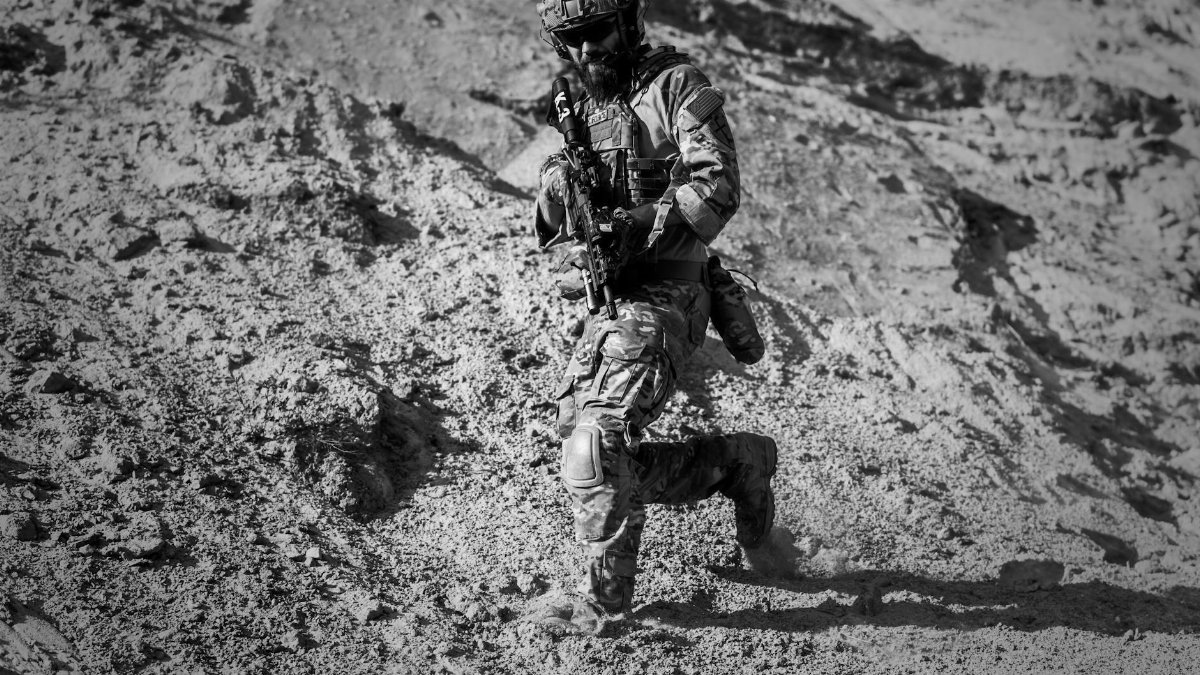 A soldier runs down a sandy hill in full combat gear under harsh desert sun.
