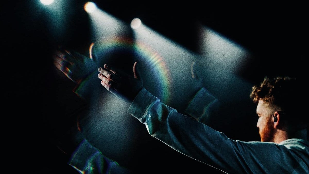 A man raises his hands in a dynamic concert performance under colorful stage lights.