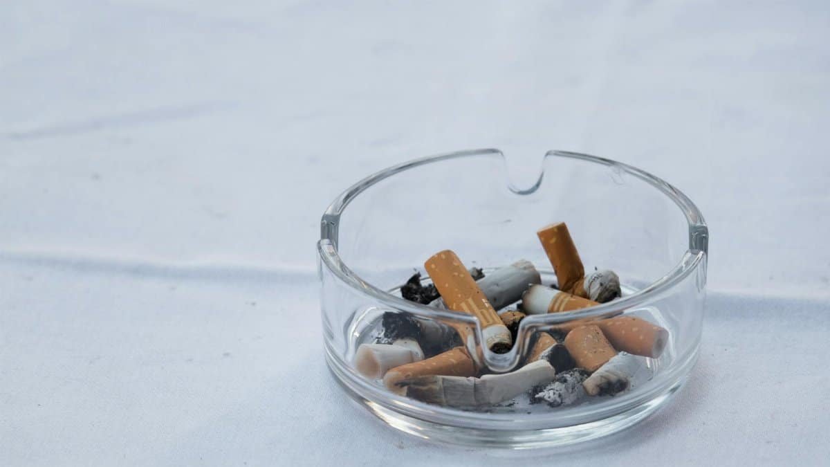 A clear glass ashtray filled with cigarette butts and ash placed on a white surface.