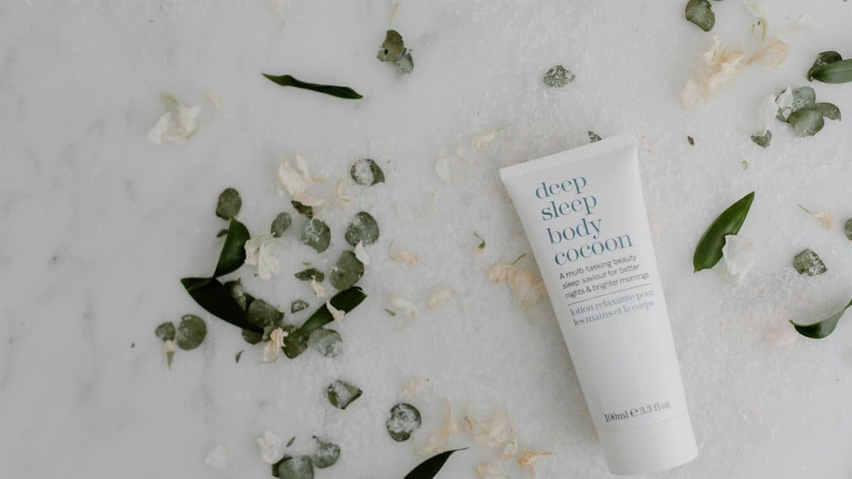 Flat lay of soothing sleep lotion surrounded by green leaves on marble, perfect for skincare themes.