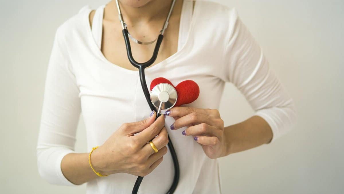 Unrecognizable female wearing white shirt while standing on white background with diaphragm of stethoscope on red handmade heart in room