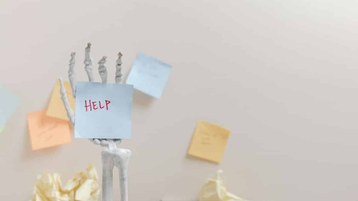 Skeleton hand holding a sticky note with 'HELP' written, against colorful notes background.