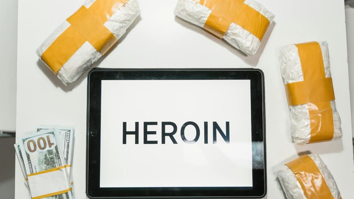 Conceptual image showing packaged heroin, money, and tablet with text in a flat lay arrangement.