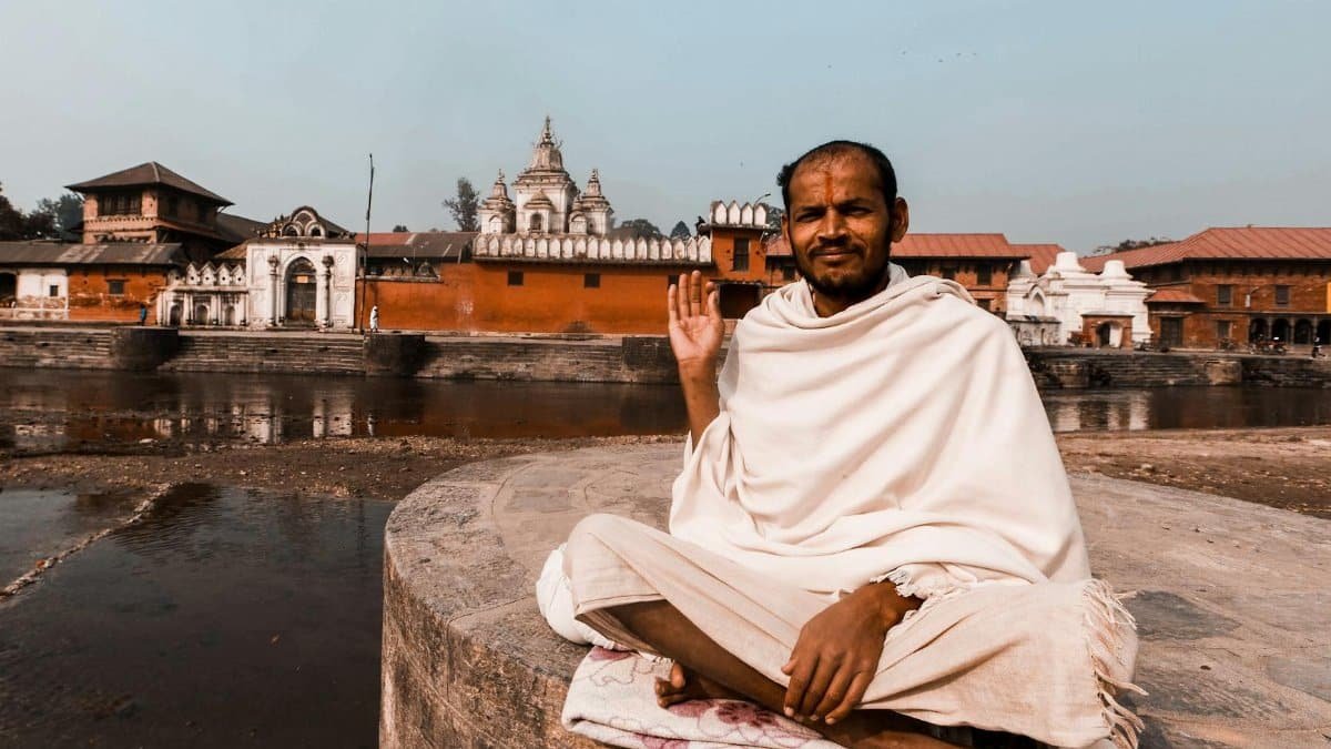 Middle-aged man in traditional attire meditating near a historic temple by a river.