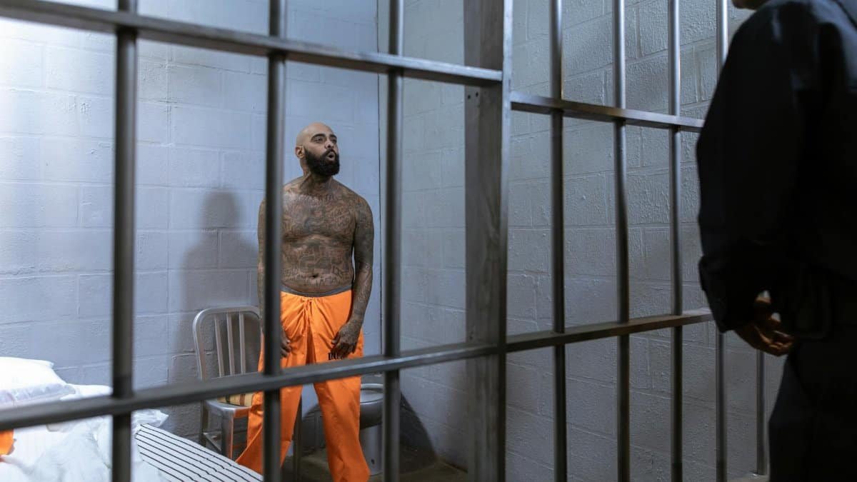 A tattooed prisoner stands behind bars in an orange jumpsuit under a guard's watch.