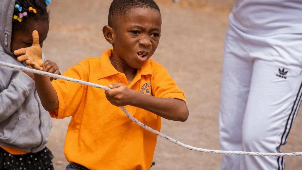 A young boy in an orange shirt enthusiastically pulls on a rope during an outdoor tug of war competition.