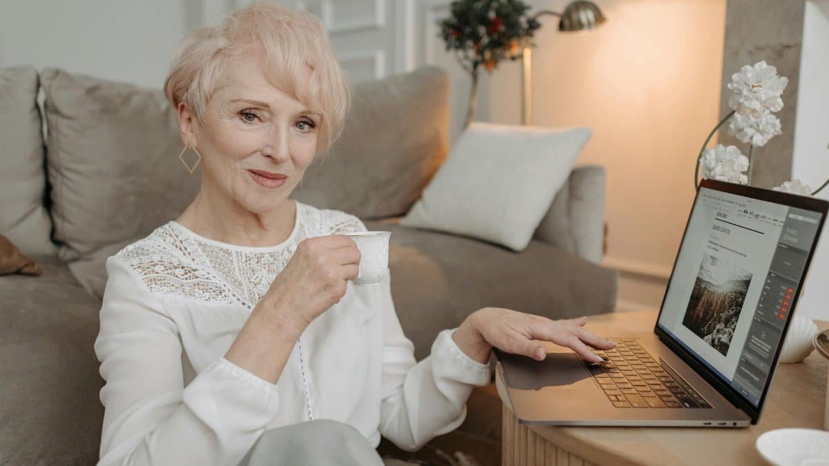 Senior woman drinking tea and working on laptop from home, epitomizing comfort in remote work.