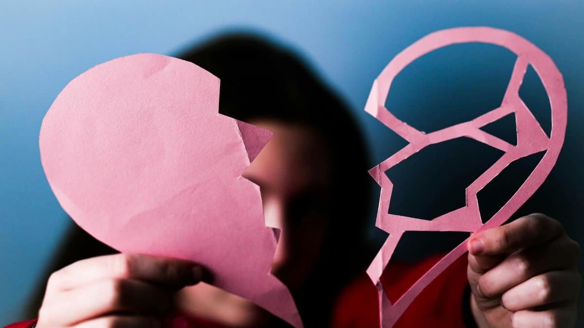Close-up of hands holding a pink broken paper heart, symbolizing love and heartbreak.