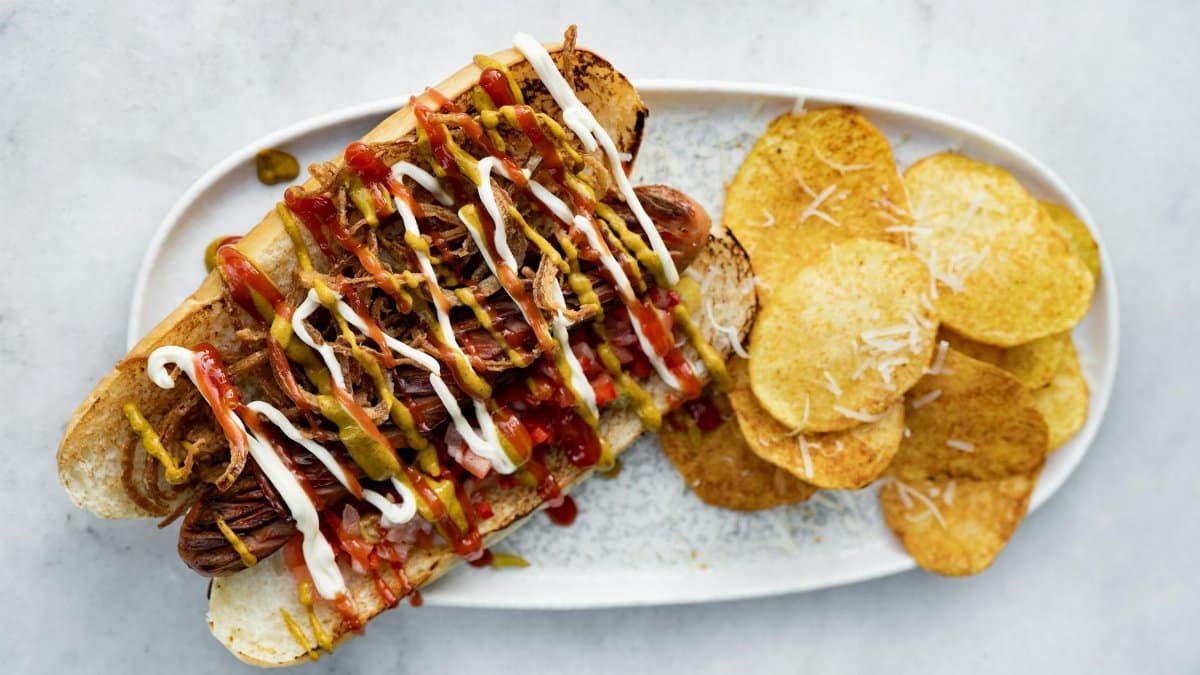 Top view of a loaded hot dog with various toppings served next to crispy potato chips on a plate.