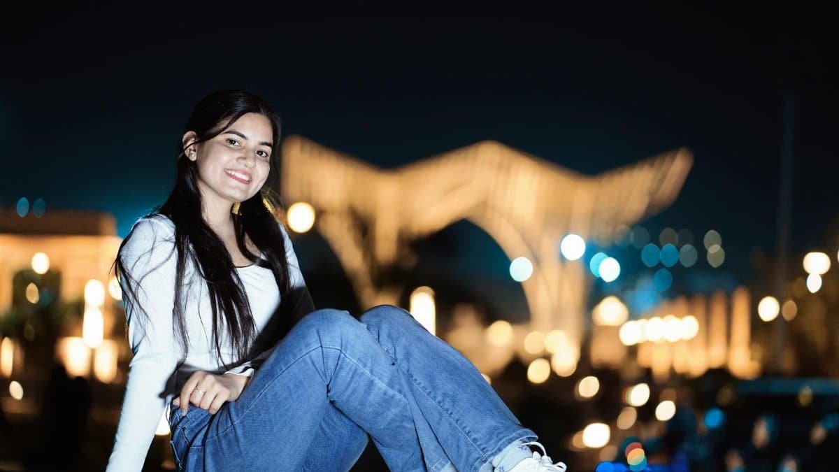 Young woman posing with a smile in an outdoor nighttime setting with blurred lights.