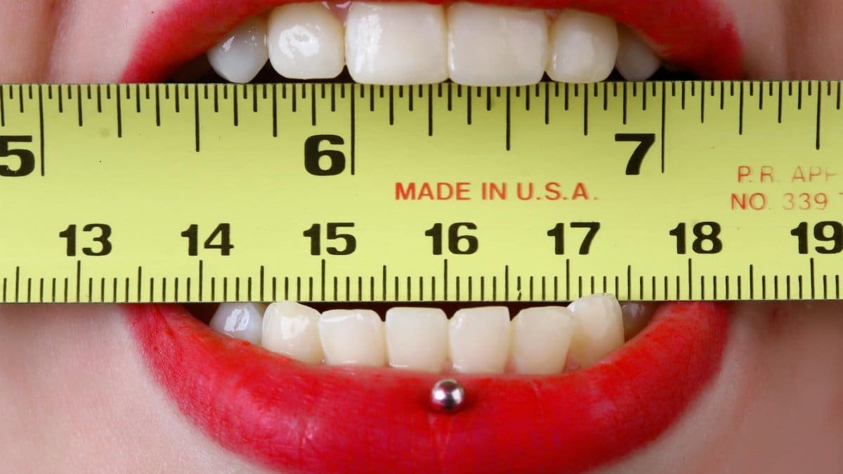 Close-up of red-lipsticked mouth holding a yellow tape measure horizontally, demonstrating measurement concept.