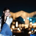 Portrait of a young woman smiling outdoors at night with city lights in the background.