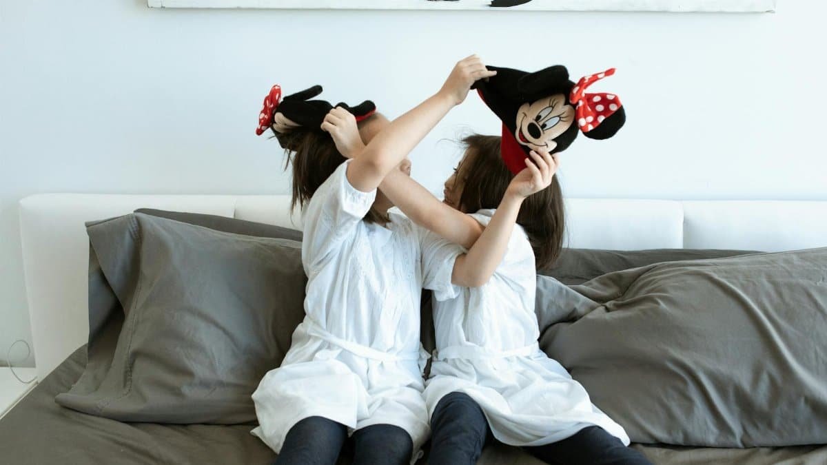 Two young girls playfully holding Minnie Mouse hats, sitting on a bed indoors.