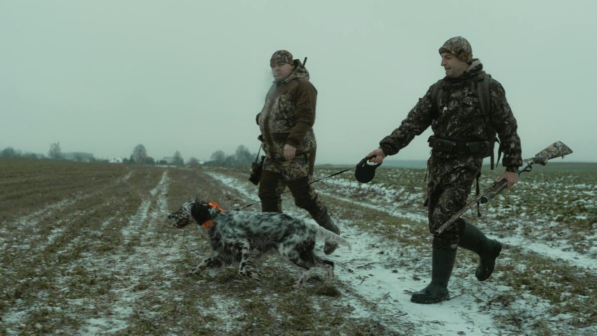 Hunters in camouflage walk with a dog in a snowy field, showcasing outdoor winter hunting expedition.