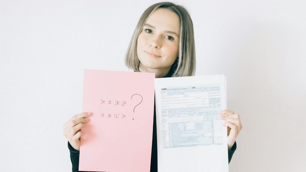 A woman in a black shirt holds tax forms and a 'Need Help?' sign, indoors.