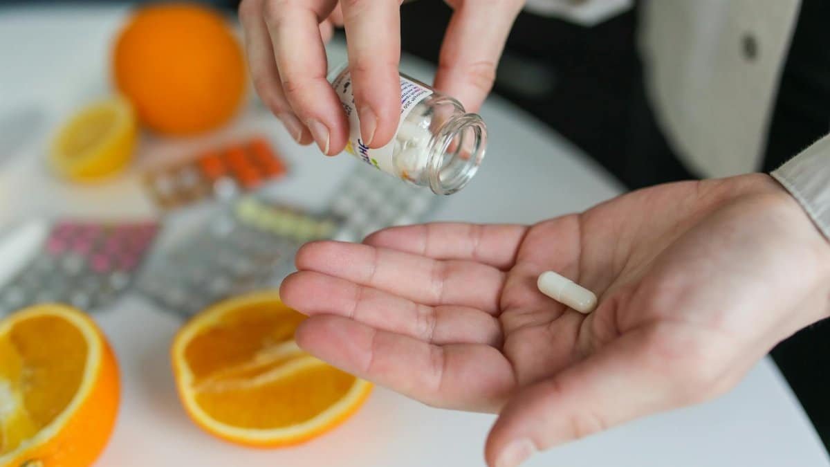 Close-up of a person taking a vitamin capsule with citrus fruit and medication on a table.