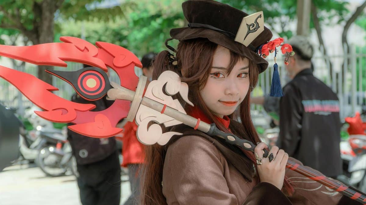 A female cosplayer dressed as Hu Tao from Genshin Impact poses outdoors with a detailed costume and weapon.