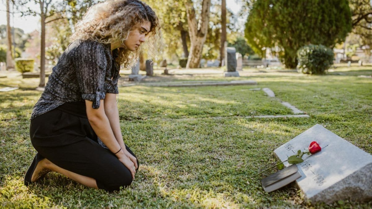 A woman kneeling by a grave in a sunlit cemetery, expressing grief and remembrance.