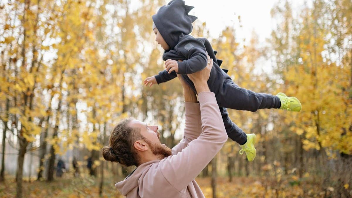 A joyful father lifting his son in a dinosaur costume amidst colorful autumn leaves in a forest.