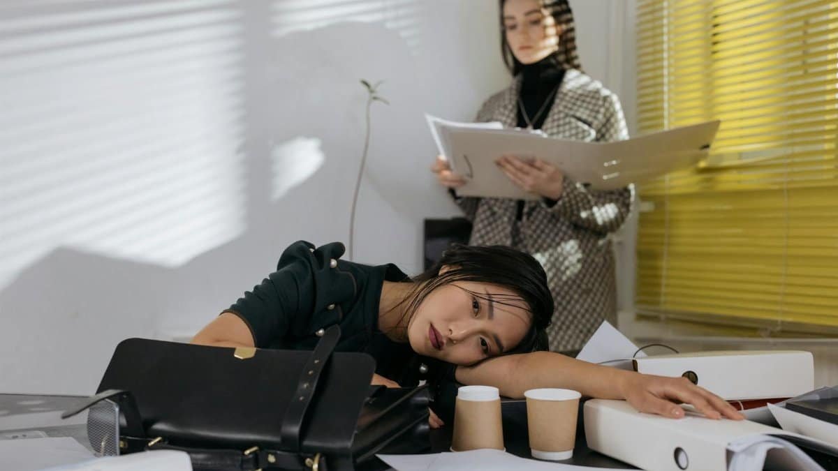 Exhausted employee resting on desk surrounded by documents, colleague in background.