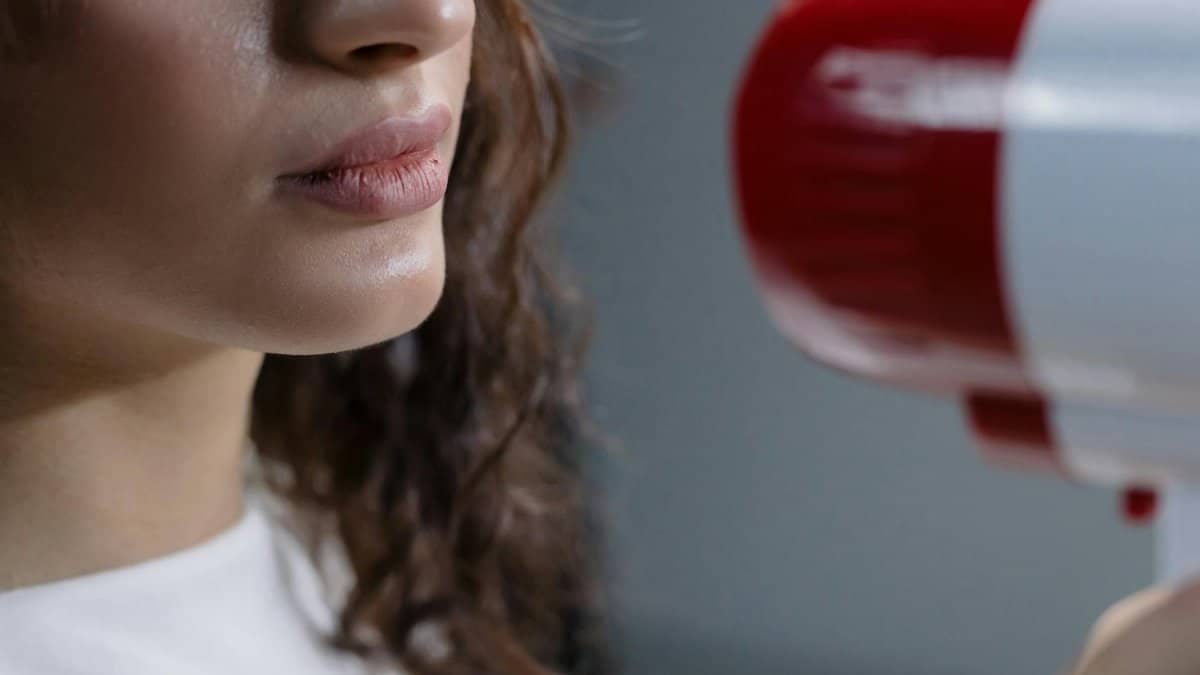 Close-up of a woman speaking into a red and white megaphone against a gray background.