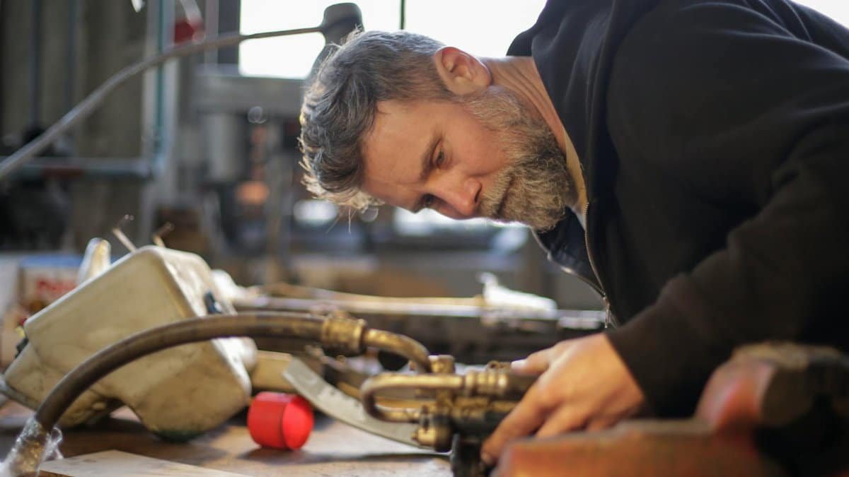 Side view of serious brutal mechanic in workwear standing at workbench and fixing metal detail while working in garage
