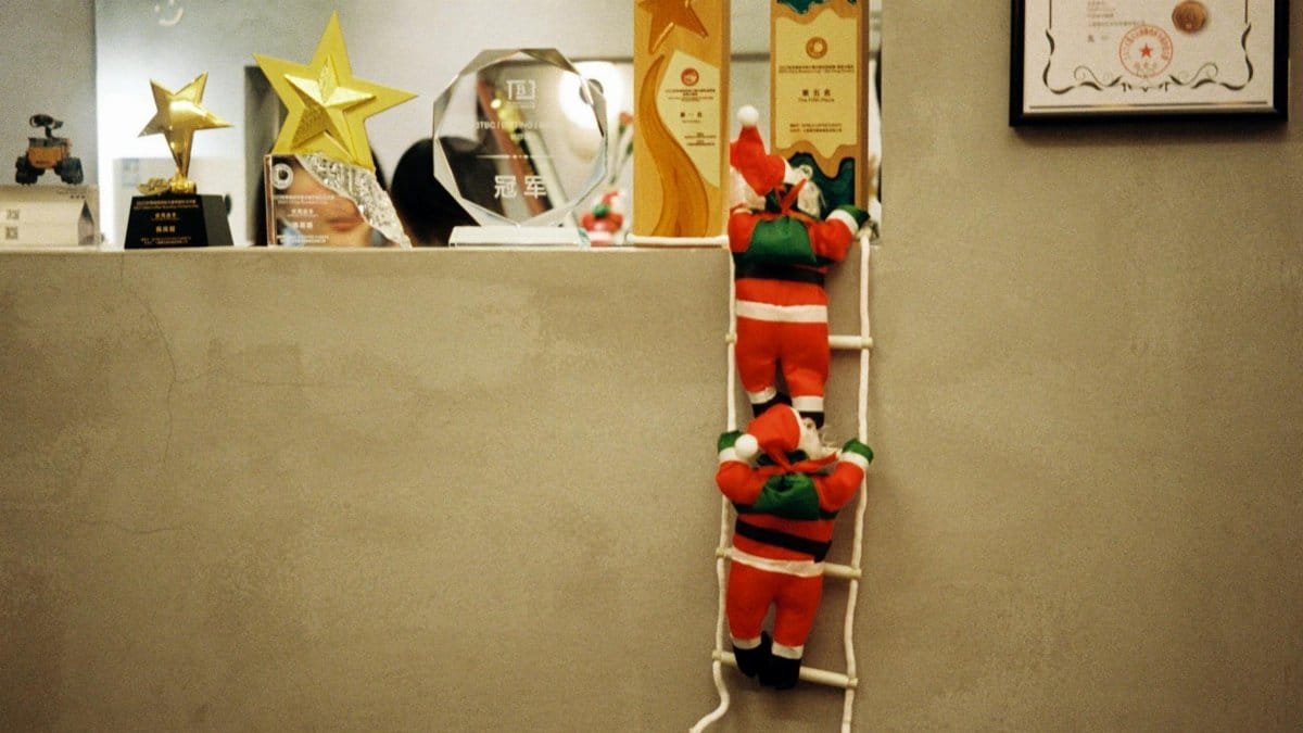 Santa Claus decorations climbing a ladder in a festive office setting, surrounded by awards and certificates.