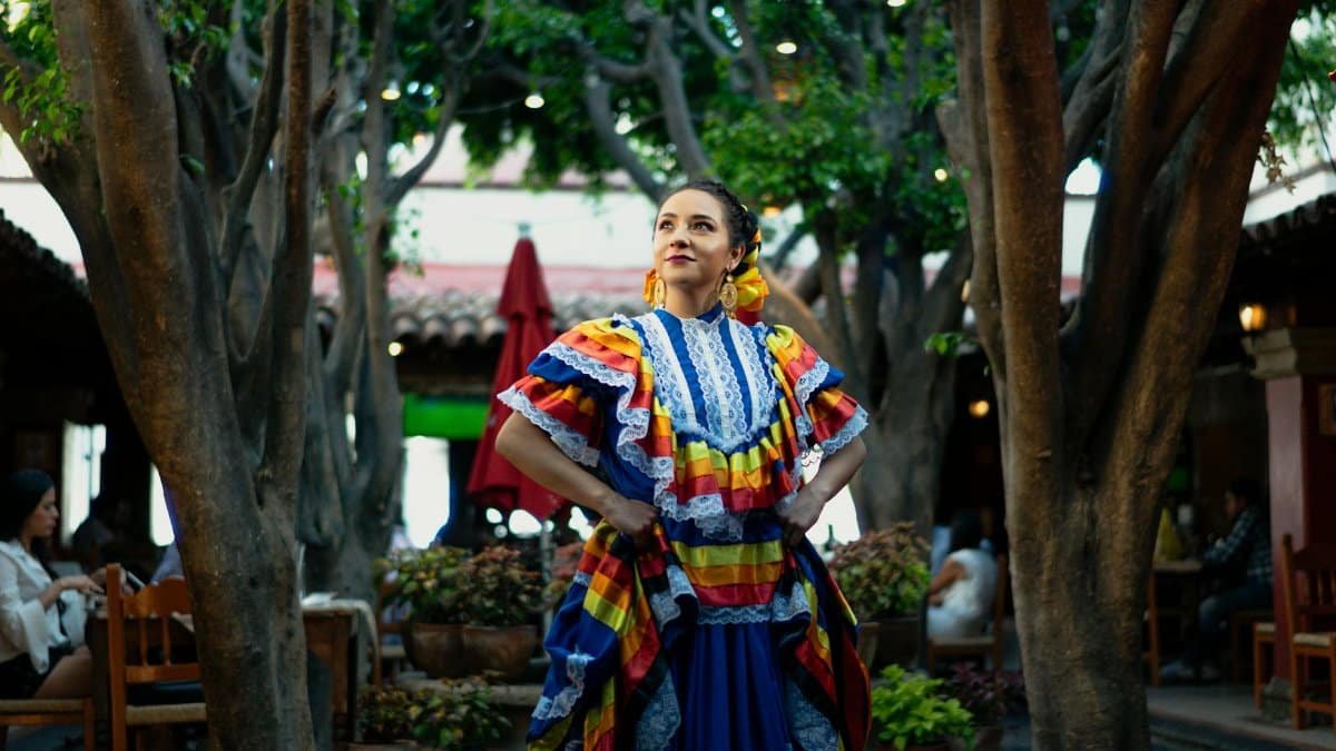 A smiling woman in traditional Mexican dress celebrates culture in a vibrant outdoor setting.