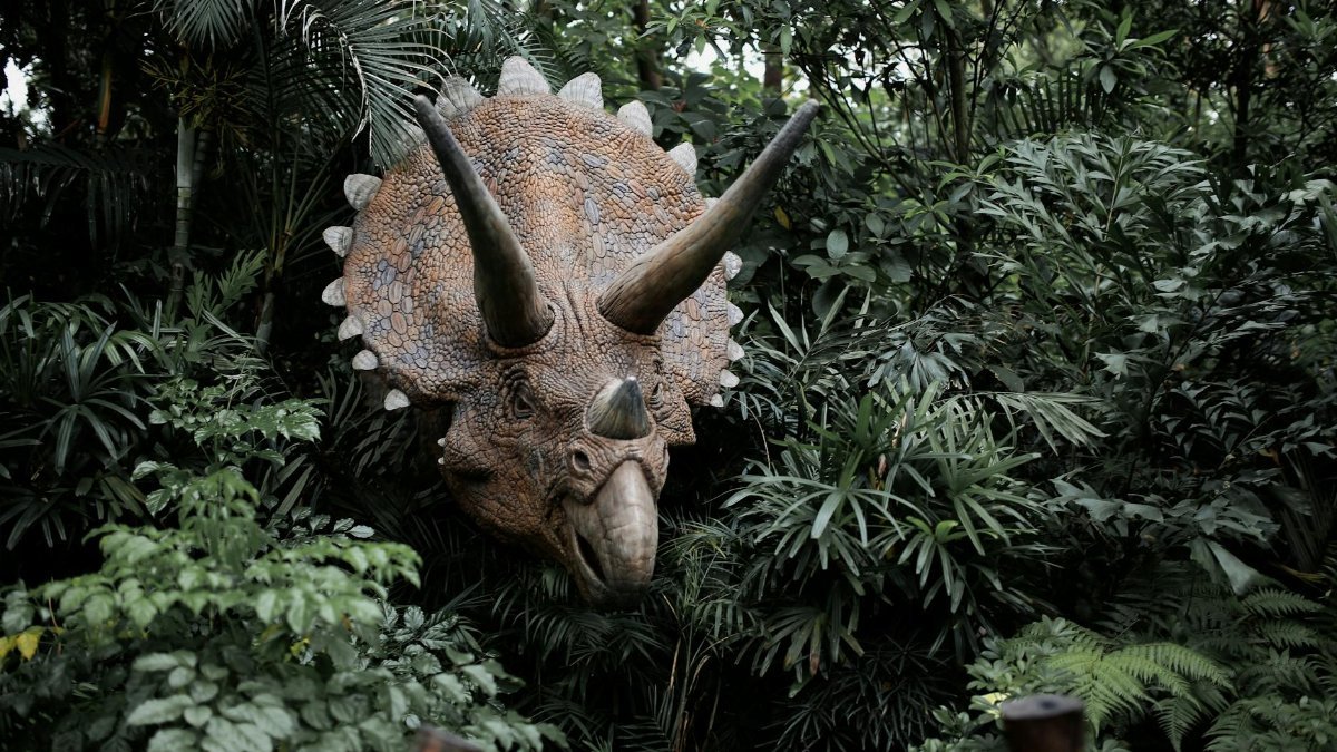 A realistic Triceratops sculpture emerges from dense jungle foliage, evoking a prehistoric atmosphere.