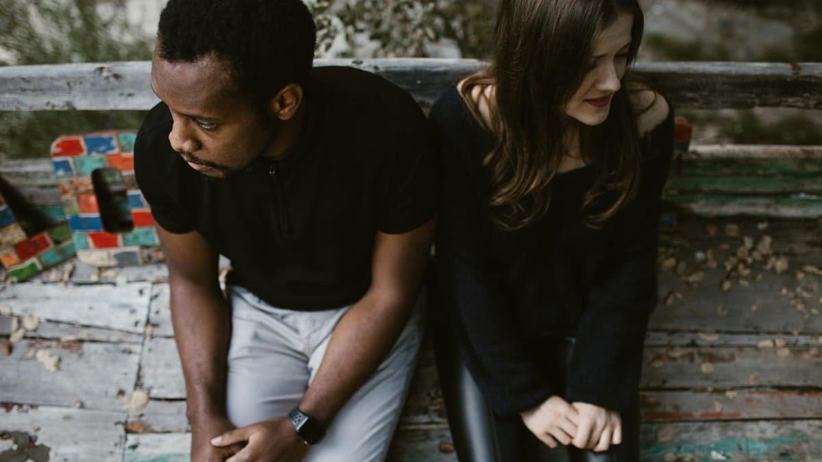 A couple sitting apart on a park bench, expressing emotions. Outdoors setting.