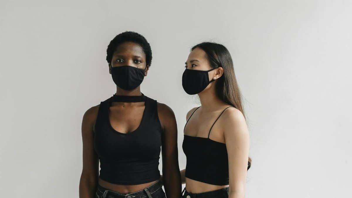 Portrait of two diverse women wearing black outfits and face masks indoors, embracing health precautions.