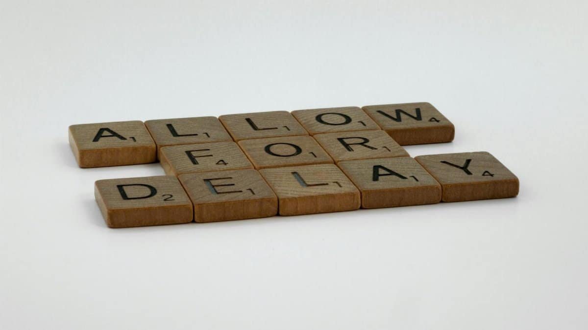 Wooden Scrabble tiles arranged on a white surface spelling 'Allow for Delay'.