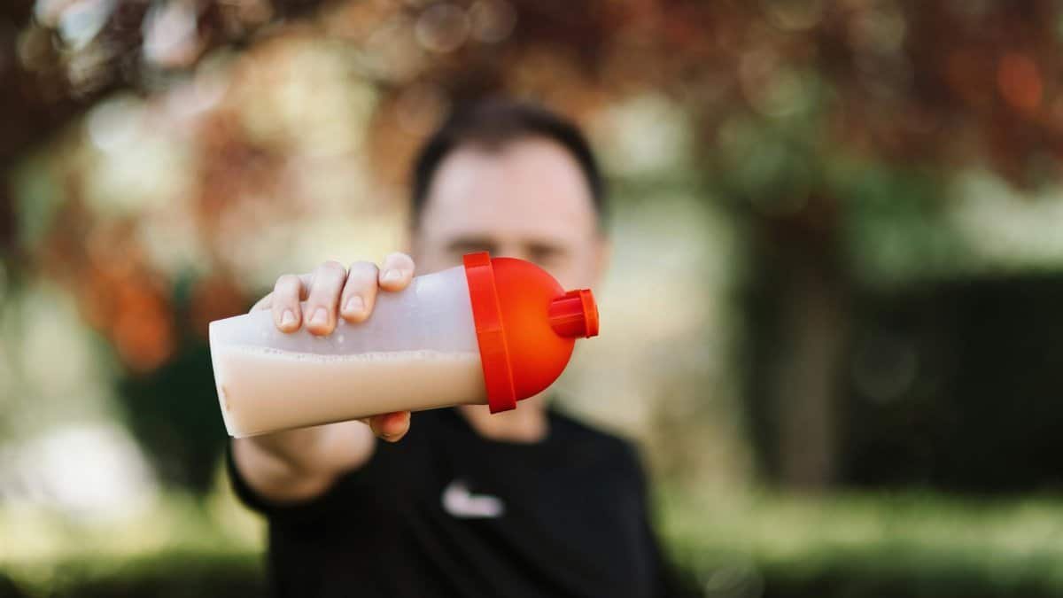 Focus on a man holding a plastic protein shake tumbler outdoors with a blurred background.