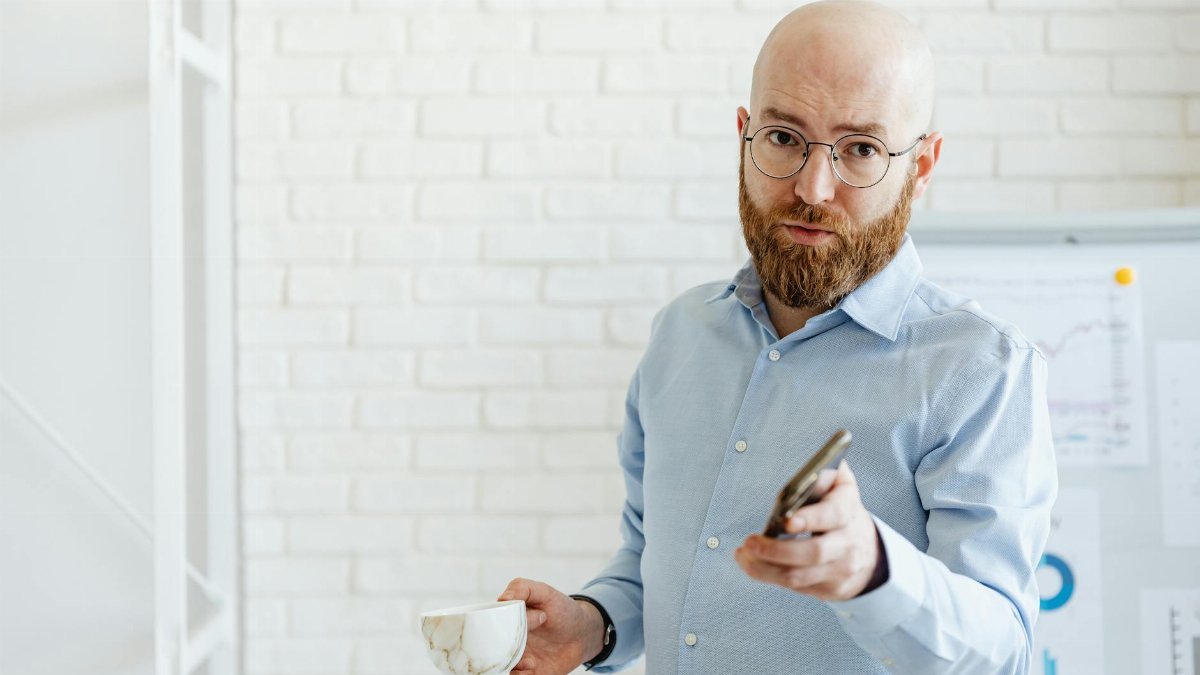 Bearded man in blue shirt holding a smartphone and cup, standing by a white brick wall.