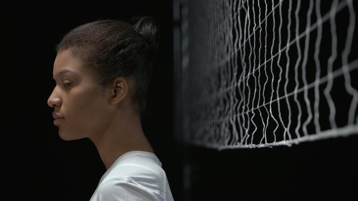 Young woman stands in profile beside a volleyball net, focused and serious.