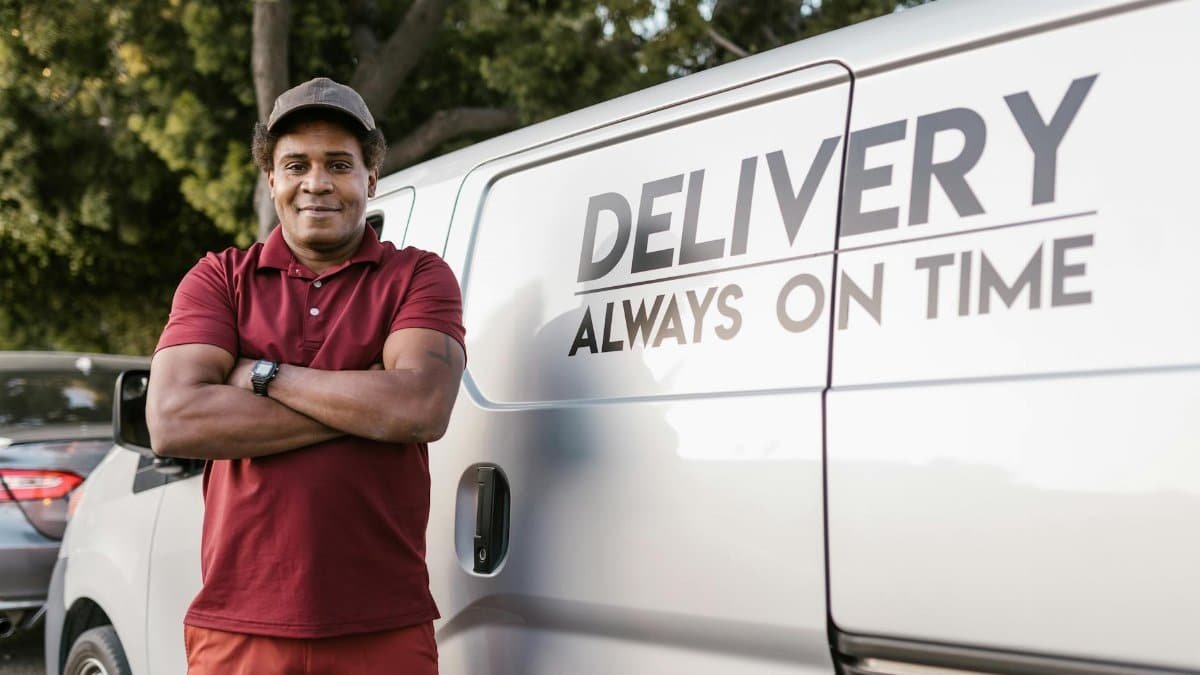 Delivery driver standing with arms crossed near a company van displaying a timely service slogan.