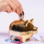A hand places a coin into a golden piggy bank surrounded by financial documents, symbolizing savings.
